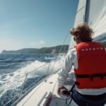 Segler auf einem Segelboot trägt eine rote Automatik-Rettungsweste, Küste im Hintergrund
