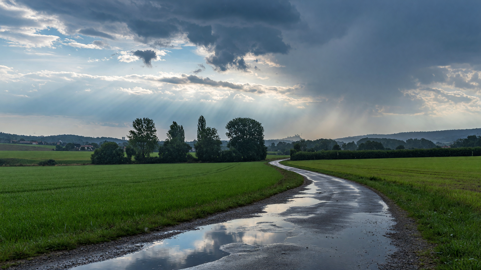 Nasse Landstraße mit Pfützen vor aufreißender Gewitterwolkendecke über grünen Feldern